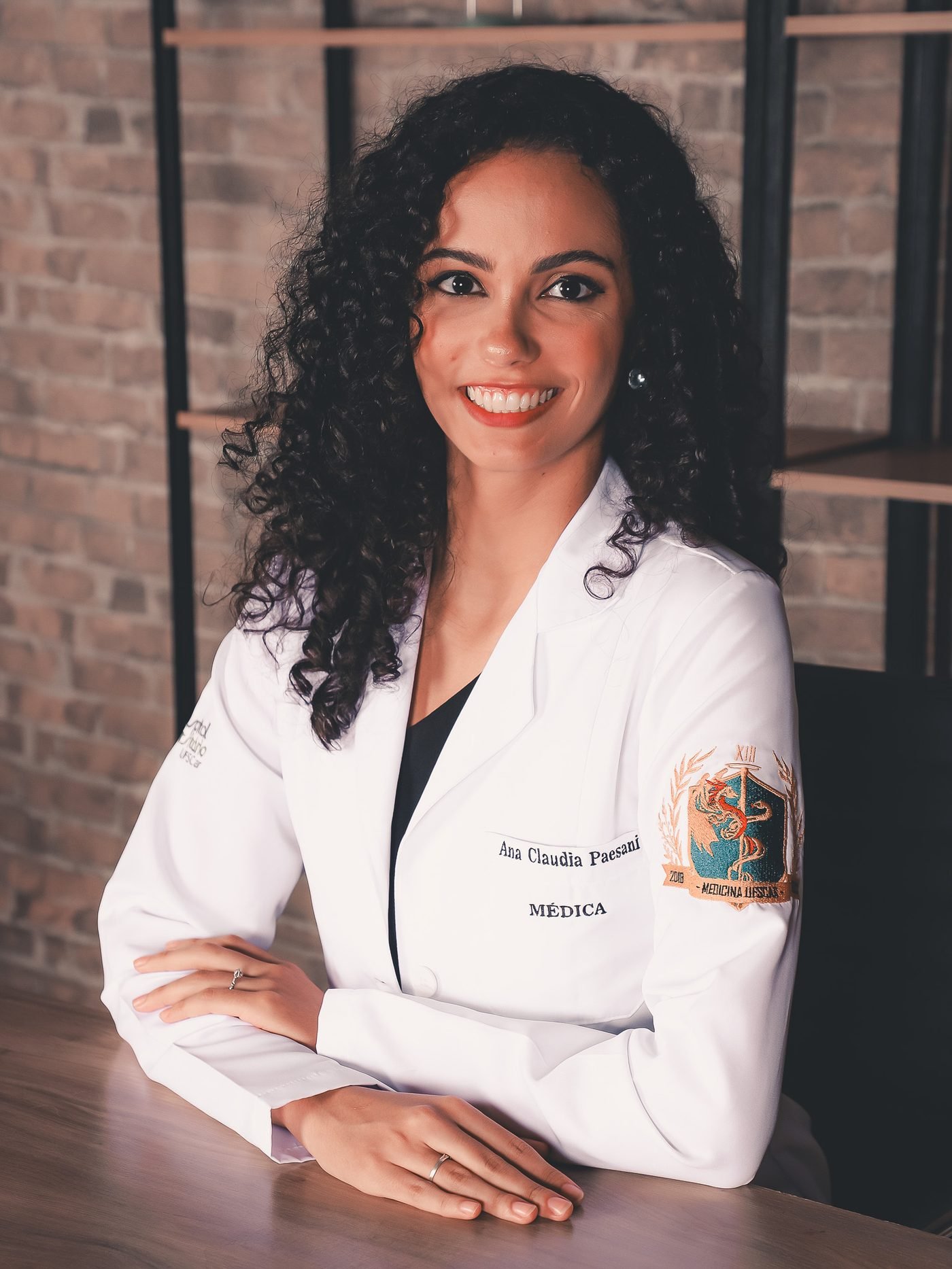 Dra. Ana Claudia sentada à mesa, sorrindo, de jaleco com o brasão da Medicina UFSCar