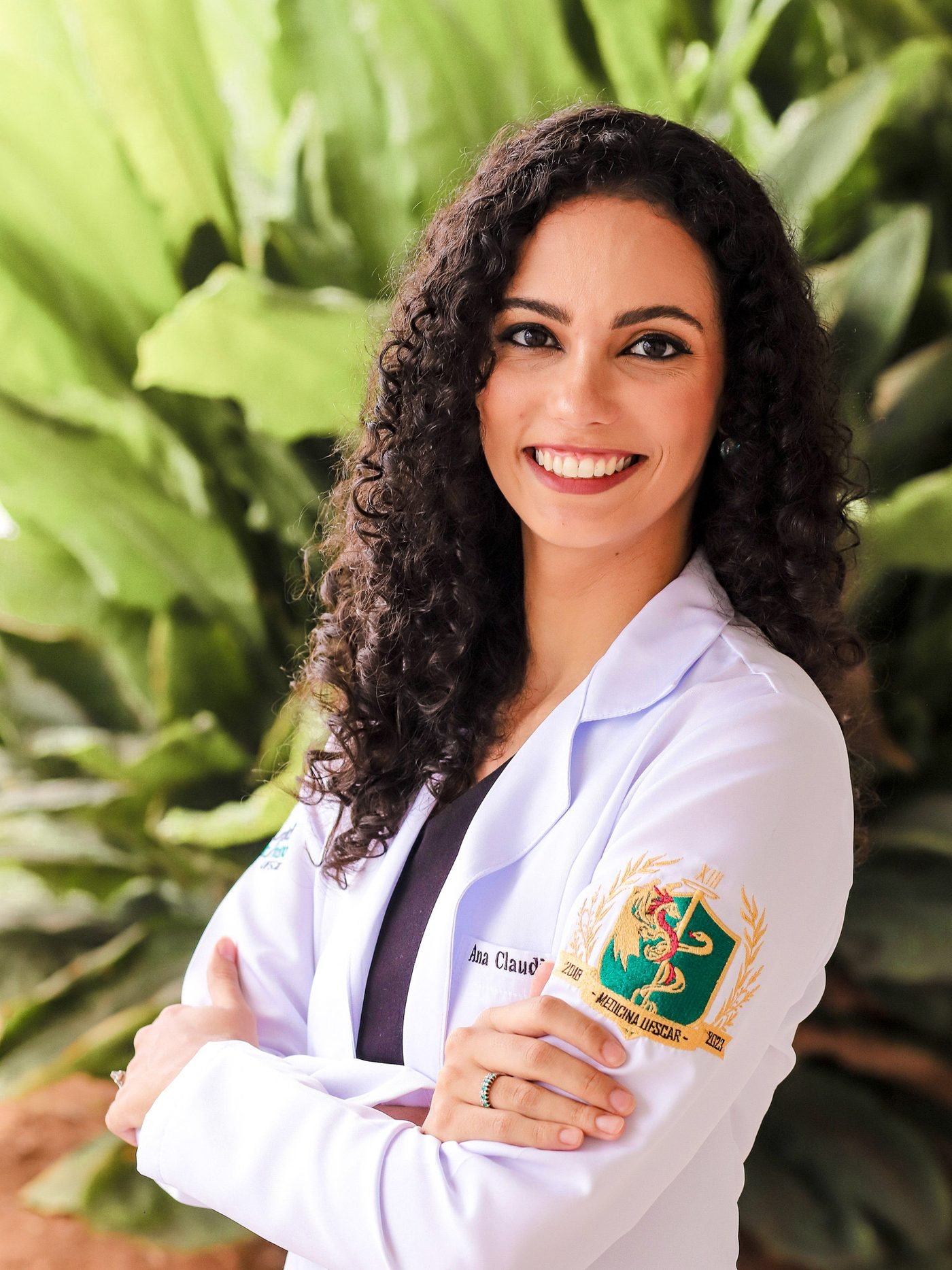 Dra. Ana Claudia Paesani sorrindo, de jaleco, em frente a plantas